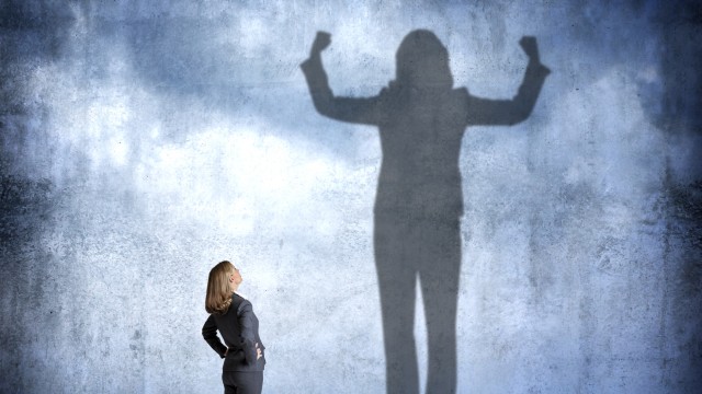 A woman stands with her hands on her hips as she looks up at her shadow that is cast on the wall in front of her that shows her flexing her muscles with power and condidence.