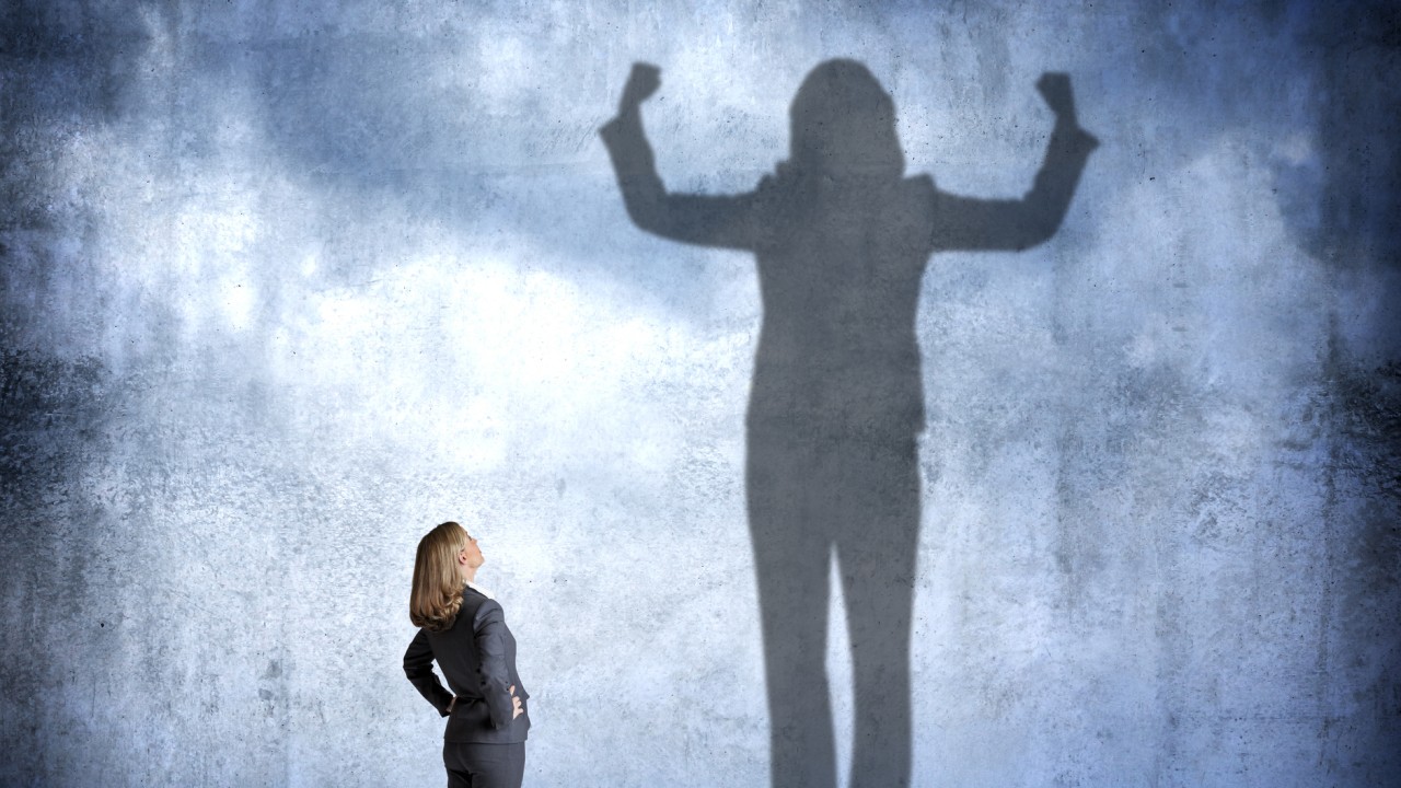 A woman stands with her hands on her hips as she looks up at her shadow that is cast on the wall in front of her that shows her flexing her muscles with power and condidence.