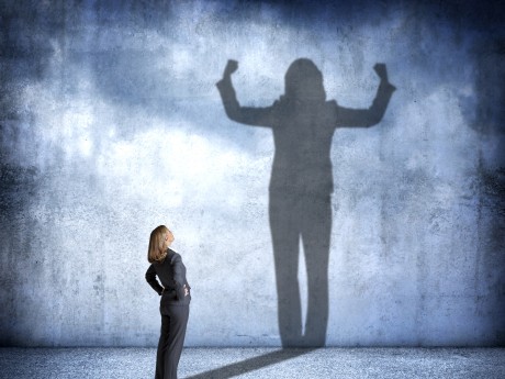 A woman stands with her hands on her hips as she looks up at her shadow that is cast on the wall in front of her that shows her flexing her muscles with power and condidence.