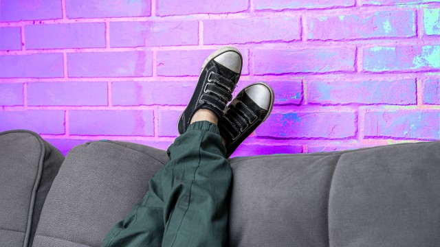 Relaxed legs of a boy in green casual pants and comfortable sneakers on a cozy sofa in the living room against a neon violet brick wall. children's feet in fashionable sneakers lie on the back of the sofa. Space for copying. Light violet neon brick background