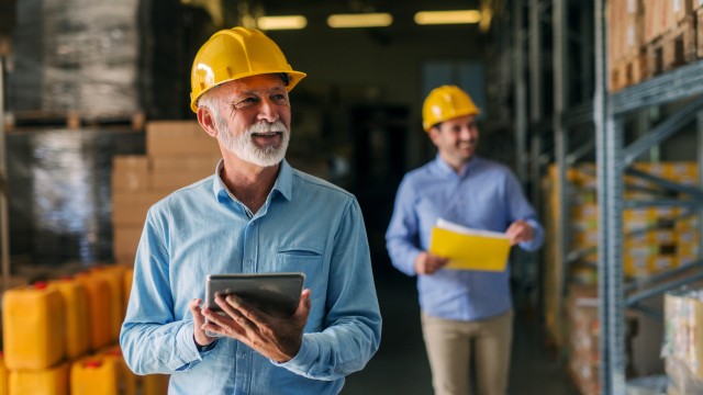 Picture of father and son walking through their warehouse with helmets on their heads. Father is holding digital tablet in his hand and looking at shelf full of boxes with smile on his face.