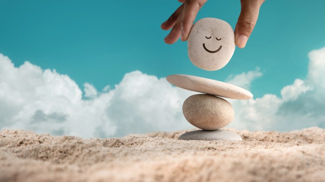 Enjoying Life Concept. Harmony and Positive Mind. Hand Setting Natural Pebble Stone with Smiling Face Cartoon to Balance on Beach Sand. Balancing Body, Mind, Soul and Spirit. Mental Health Practice